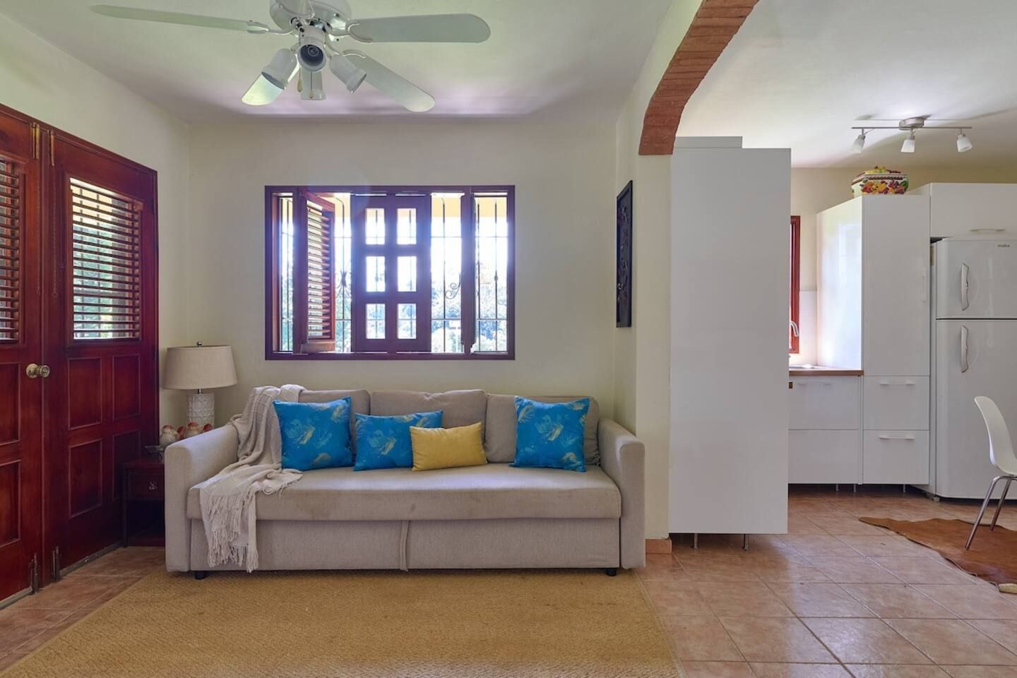 Un espacio de estar con un sofá, un ventilador de techo, una ventana con persianas y una cocina con refrigerador.