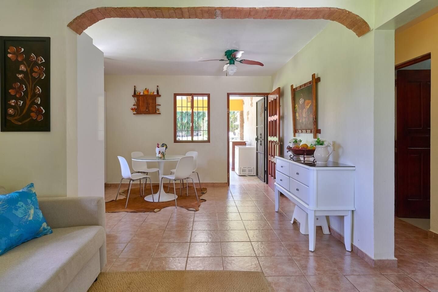 Un coin repas avec une table ronde et des chaises, un ventilateur de plafond, et une vue sur une pièce lumineuse, accessible par une porte ouverte.