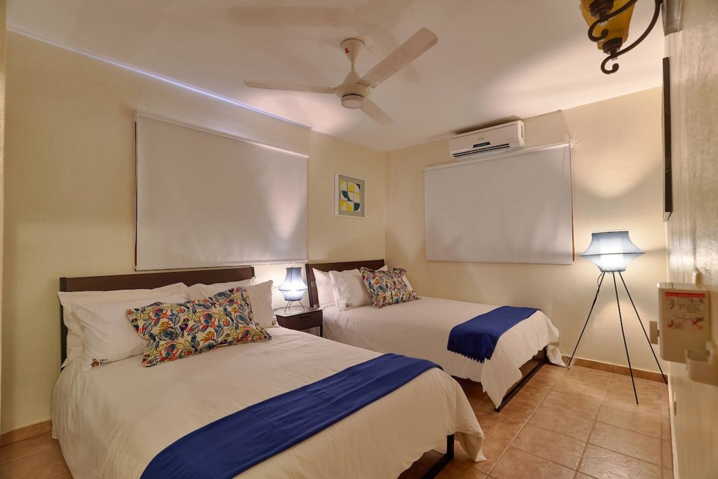 Habitación de hotel con dos camas, ventilador de techo, unidad de aire acondicionado, lámpara de pie y ambientador de pared.