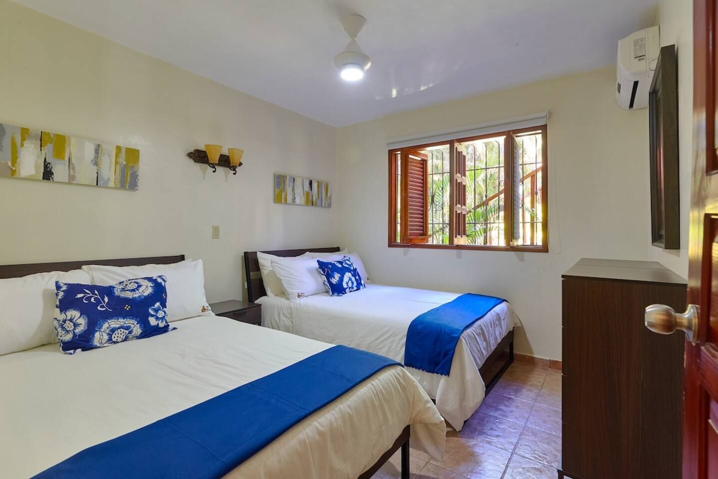Una habitación con dos camas, ventilador de techo, ventana con contraventanas y aire acondicionado de pared.