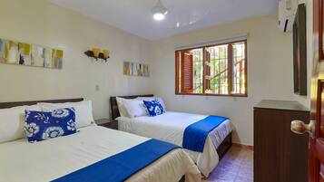 Una habitación con dos camas, ventilador de techo, ventana con contraventanas y aire acondicionado de pared.