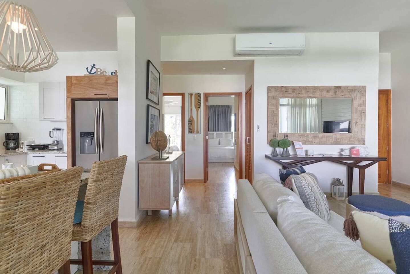 A modern living room with a sofa, a dining area with wicker chairs, a kitchen with a refrigerator, and a hallway leading to a bedroom.