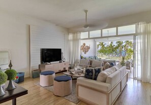 A spacious living room with a large flat-screen TV, a sofa set, and a coffee table. There is a balcony with a view of palm trees. - ®{The~Palms~View}@PlayaBonita +Beachfront+Pool+BBQ (Las Terrenas)