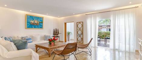 A spacious living room with a white sofa, a wooden coffee table, and a dining area with wicker chairs. A large painting and decorative ship models adorn the walls.