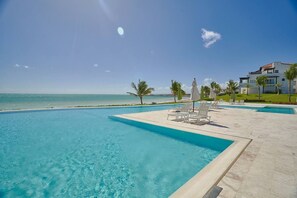 Piscina azul translúcida com espreguiçadeiras e guarda-sóis, vista para a praia e um edifício moderno ao fundo.