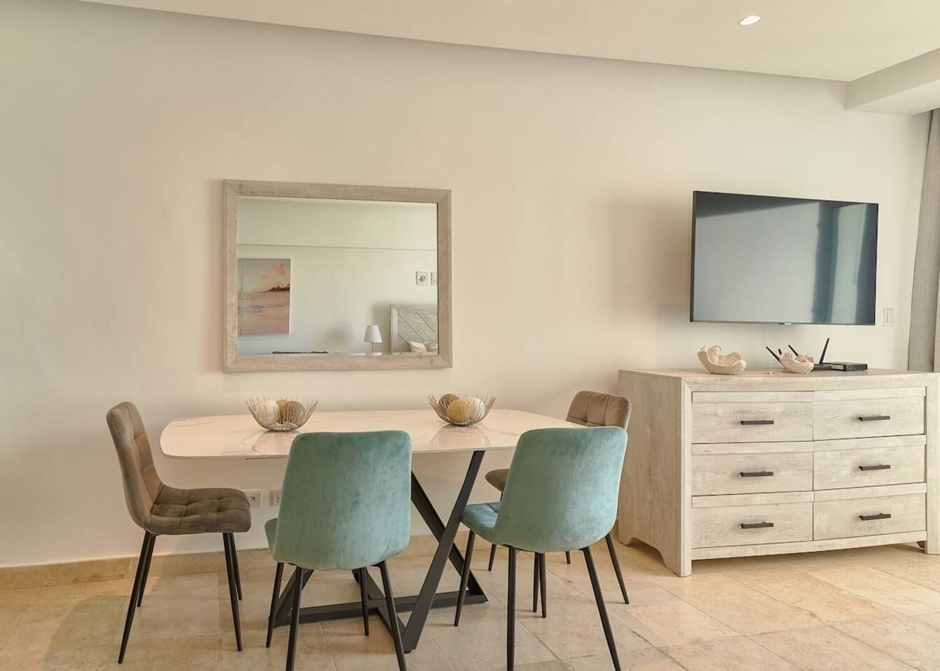 Uma sala de jantar com uma mesa branca, quatro cadeiras, um espelho grande e uma TV em um móvel.