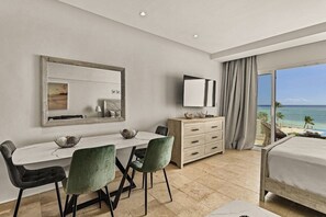 Ett hotellrum med en säng, ett matbord med stolar, en tv, en byrå och ett stort fönster med utsikt över stranden.