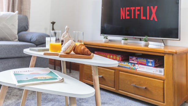 A living room with a television displaying 'NETFLIX,' a small table with a magazine, a glass of orange juice, and a croissant.