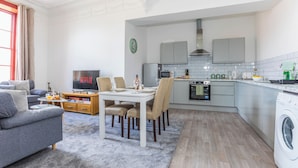 A modern living room with a sofa, TV, and dining area. A kitchen with appliances and a washing machine is visible.
