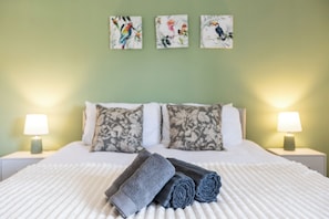 A neatly made bed with rolled towels, two nightstands with lamps, and three wall-mounted bird paintings.