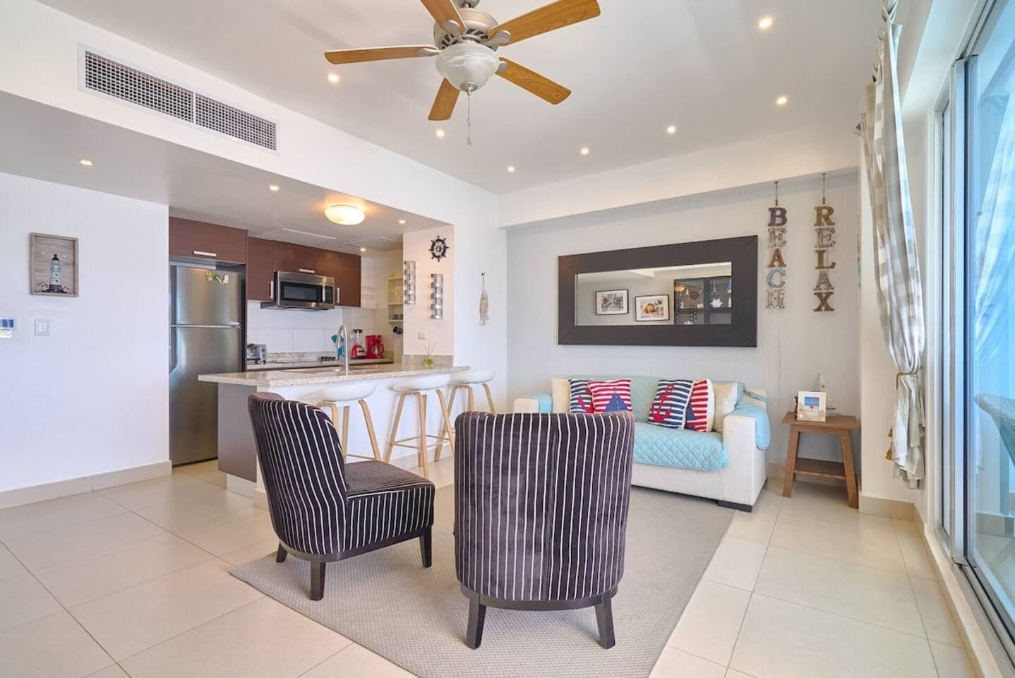 A modern living room with a sofa, two striped chairs, a kitchen area, and a ceiling fan.