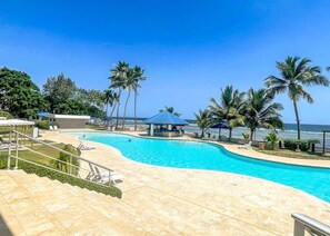 A swimming pool with a curved design, surrounded by palm trees and lounge chairs, with a view of the ocean. - ®{Las~Olas~Beach~Condo}@JuanDolio +BeachFront+Pool (Juan Dolio)