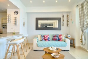 A modern living room with a sofa, a coffee table, and a kitchen area in the background. - ®{Las~Olas~Beach~Condo}@JuanDolio +BeachFront+Pool (Juan Dolio)