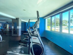 A gym with treadmills and exercise equipment, large windows with a view of palm trees, and a blue wall. - ®{Las~Olas~Beach~Condo}@JuanDolio +BeachFront+Pool (Juan Dolio)