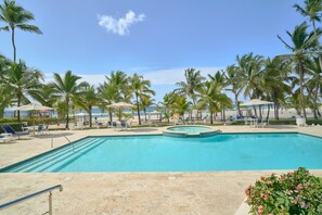 A swimming pool with lounge chairs and umbrellas, surrounded by palm trees and a beach view. - ®{Costa~Sol~Views} @JuanDolio +Pool+Beachfront (Playa Juan Dolio)