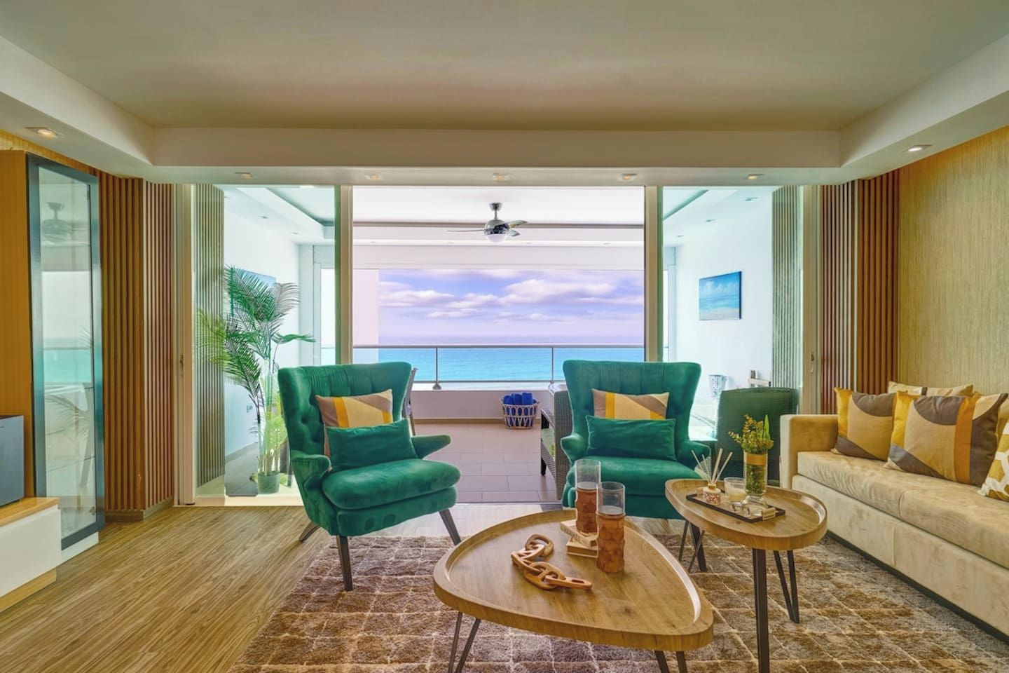 A modern living room with two green armchairs, a beige sofa, and a coffee table. The room has a large glass door leading to a balcony with a view of the ocean.