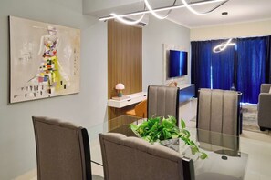 A modern dining area with a glass table, grey upholstered chairs, a decorative plant centerpiece, a flat-screen TV mounted on the wall, and a blue curtain in the background. - ® 2BR{Ajega~Views} @Downtown SDQ +Pool+Gym (Santo Domingo)