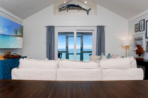 A living room with a white sofa, a wooden floor, a large window with a view of the sea, and a mounted fish on the wall. - My Tide (Port St. Joe)