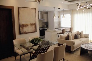 A modern living room with a glass dining table, beige sofas, and a kitchen in the background. - ®2BR{Santorini~Views} @Downtown SDQ +Lounge+Gym (Santo Domingo)