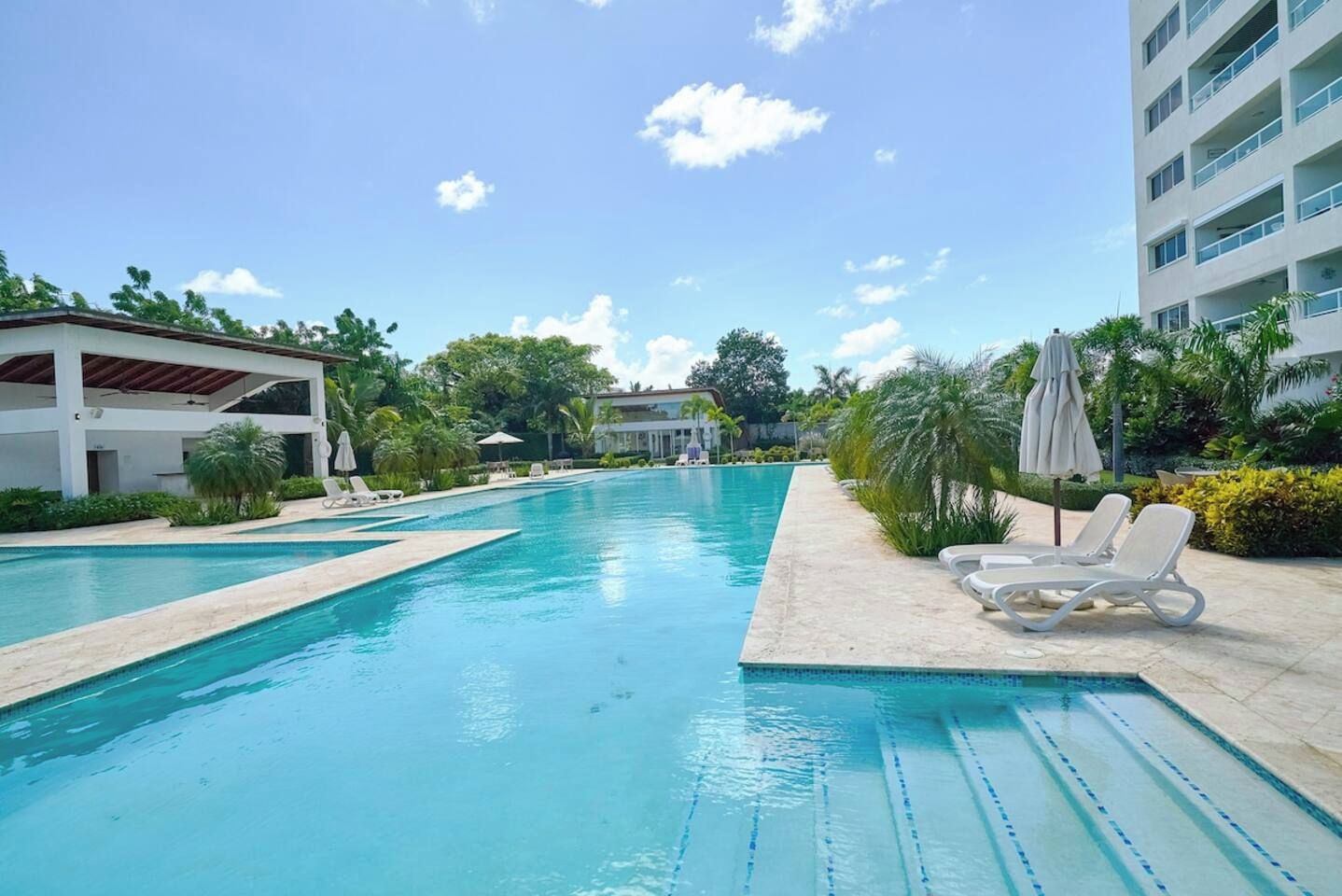 Piscina com espreguiçadeiras e guarda-sóis, rodeada por vegetação e edifícios.