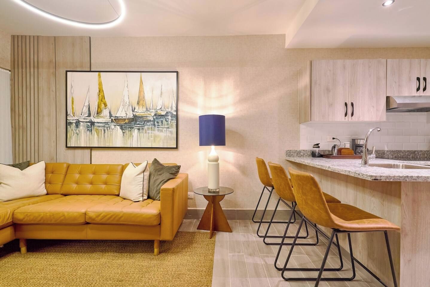 Een moderne woonkamer met een bank, een schilderij, een lamp en een keukenhoek met een spoelbak en stoelen.