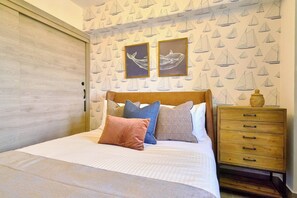 Una camera da letto con un letto, cuscini, una cassettiera in legno e una parete con carta da parati a tema vela.