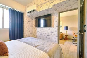 A hotel room with a bed, a flat-screen TV mounted on the wall, a window with curtains, and a door leading to a dining area. - ®{Coral Cliffs~Executive} @JuanDolio + Pool + Gym (Juan Dolio)