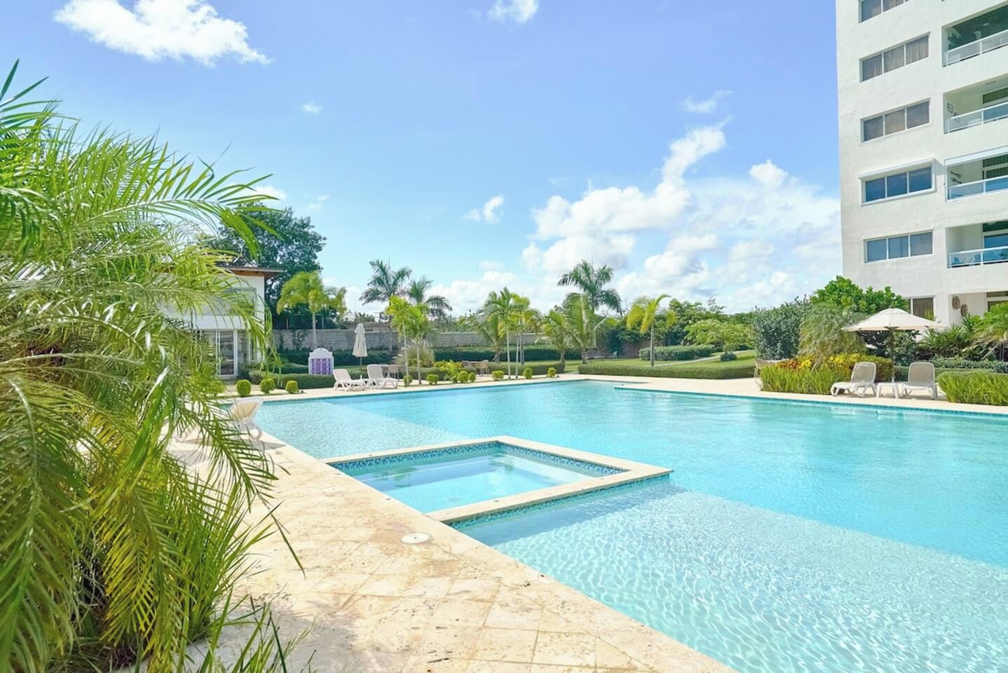 Une piscine aux eaux cristallines, bordée de chaises longues et de palmiers, le tout entouré d’un immeuble à plusieurs étages.