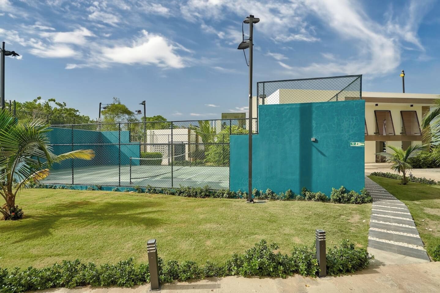 A tennis court with a fence, a pathway, and a building with a green wall.