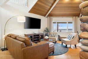 A living room with a brown leather sofa, a beige armchair, a coffee table, a flat-screen TV, and a large window with a view. - Point Whitehorn Cottage by Gather: Coastal Retreat w/ Ocean Views, Deck & Beach (Blaine)