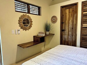 Une chambre à coucher avec un lit, une porte en bois, une étagère fixée au mur ornée d’objets décoratifs et une fenêtre avec des stores.