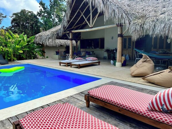 Un espace piscine avec des chaises longues et une structure dotée d’un toit de chaume.