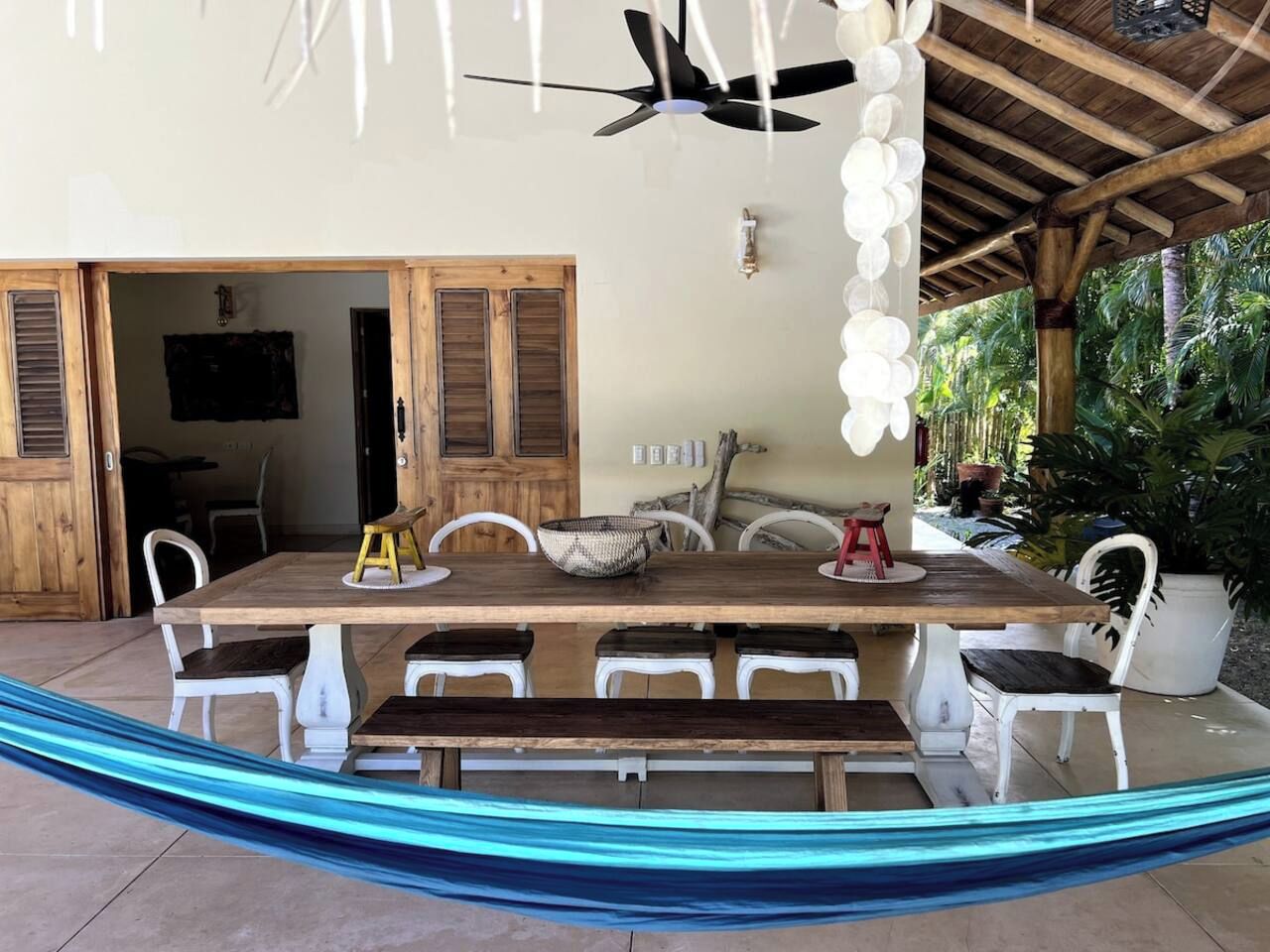 Uma área de refeições ao ar livre coberta, com uma mesa de madeira, cadeiras brancas, uma rede e um ventilador de teto.