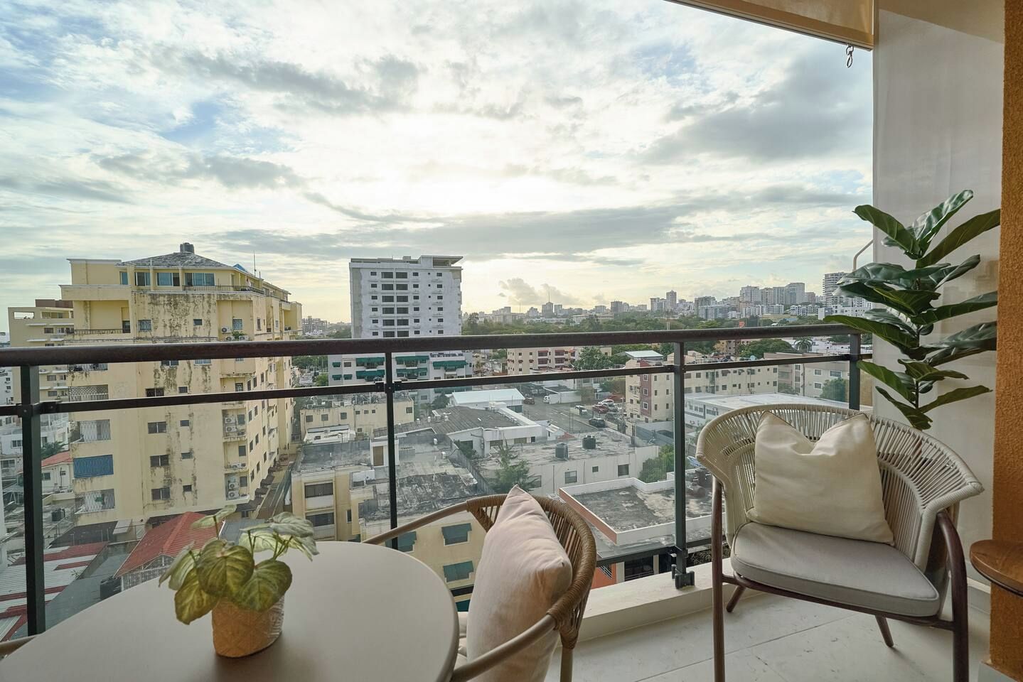 A balcony with a view of a cityscape, a table, two chairs, and a potted plant.