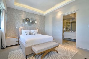 Une chambre à coucher moderne avec un lit, des tables de chevet, un banc, une œuvre d’art murale et une salle de bain visible à travers une porte ouverte.