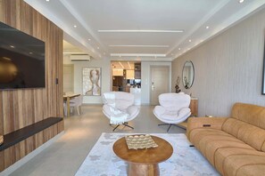 A modern living room with a flat-screen TV, two white armchairs, a beige sofa, and a wooden coffee table. - 3BR Elegant Spot @DowntownSD +Family+Pool (Santo Domingo)