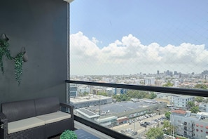 A balcony with a view of a cityscape, a grey sofa, and hanging plants. - 3BR Elegant Spot @DowntownSD +Family+Pool (Santo Domingo)