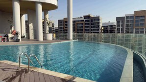 Una piscina con barrera de vidrio, rodeada de edificios modernos y una zona de estar con mesas y sillas.