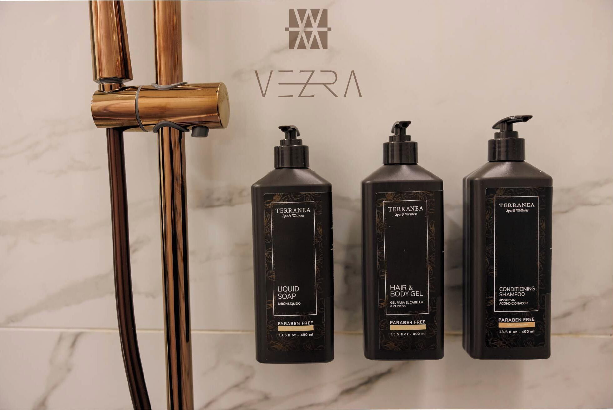 Three black bottles labeled TERRANEA Spa & Wellness, including LIQUID SOAP, HAIR & BODY GEL, and CONDITIONING SHAMPOO, next to a shower with a brass faucet.