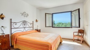 Ein Schlafzimmer mit einem Bett, einem Sessel, einem Nachttisch, einer Lampe, Wandleuchte und einem Fenster mit Blick.