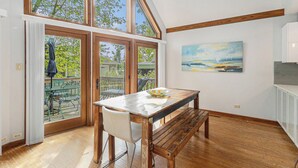Uma área de refeições com mesa de madeira e bancos, janelas amplas e um quadro na parede.
