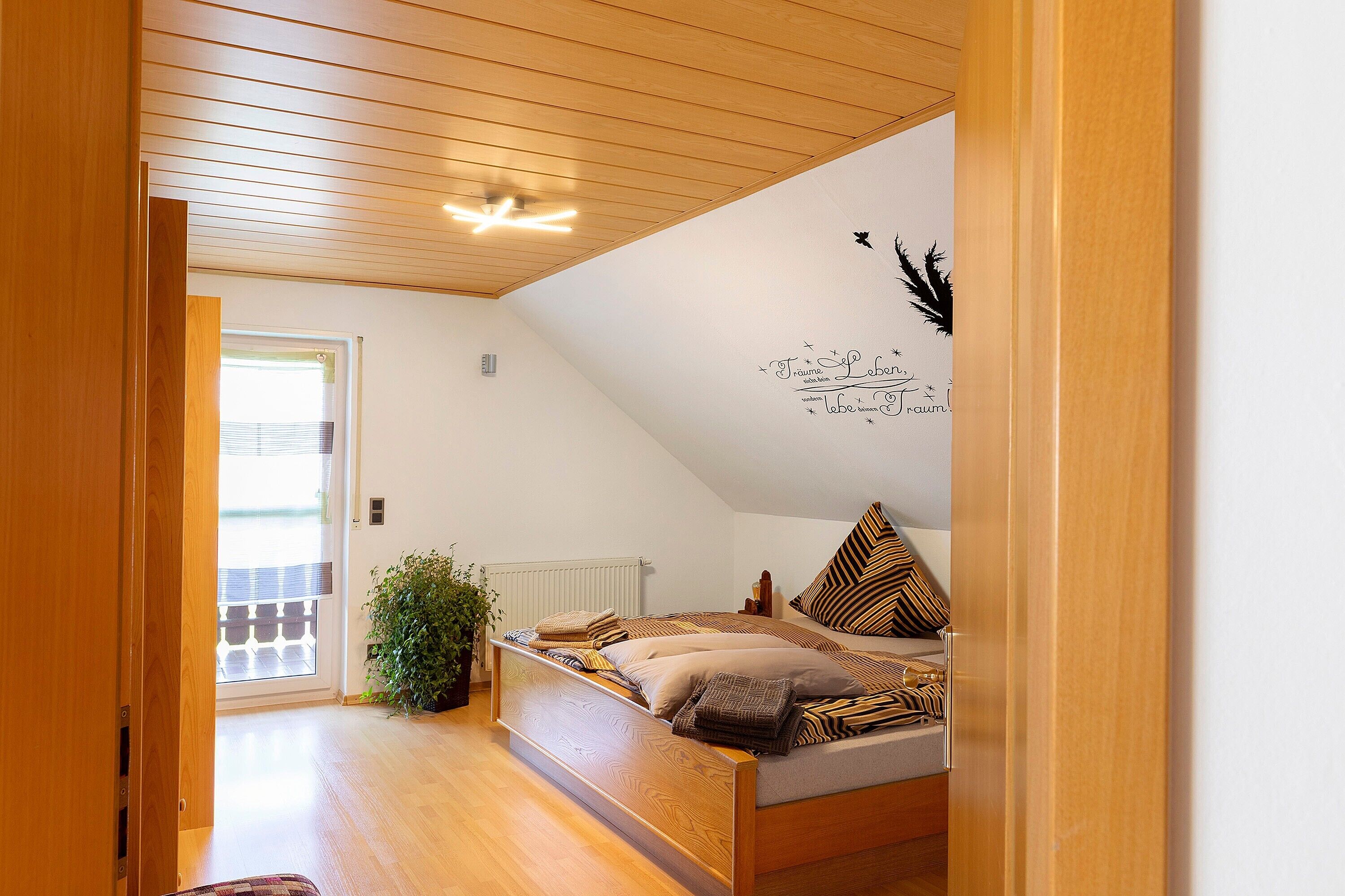 Une chambre avec un plafond en pente, un lit avec une literie à motifs, un radiateur et une plante en pot.