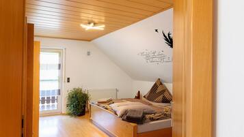 Une chambre avec un plafond en pente, un lit avec une literie à motifs, un radiateur et une plante en pot.