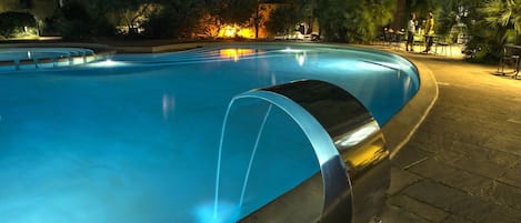 A swimming pool with a curved edge, surrounded by a paved walkway and trees.