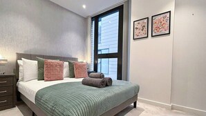 Ein Schlafzimmer mit einem Bett, Kissen, einem Nachttisch, einem Fenster und zwei gerahmten Bildern an der Wand.