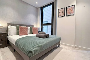 Un dormitorio con una cama, almohadas, una mesita de noche, un ventanal y dos cuadros en la pared.