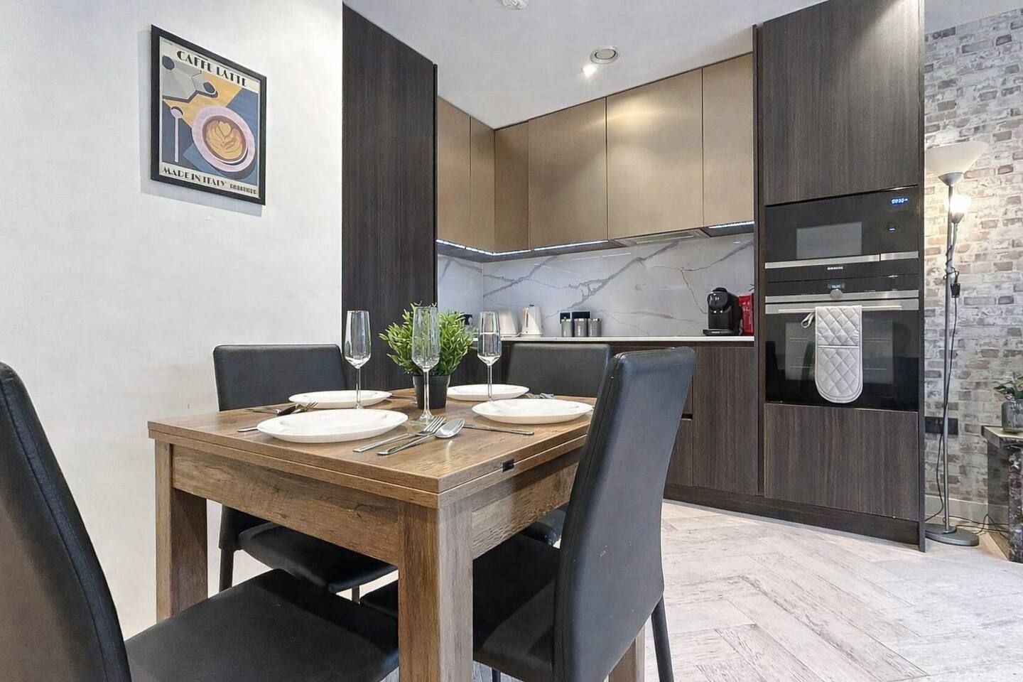 Un'area pranzo moderna con un tavolo in legno apparecchiato per quattro, una cucina con mobili scuri e una parete di mosaico in marmo, e un muro a mattoni a contrasto.