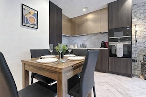 Un comedor moderno con una mesa de madera dispuesta para cuatro, una cocina con armarios oscuros y un salpicadero de mármol, y una pared de ladrillo decorativa.