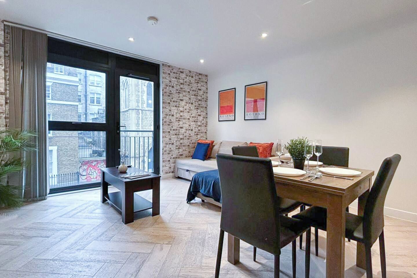 A modern dining area with a wooden table set for two, a sofa, and a large window with a view of buildings.
