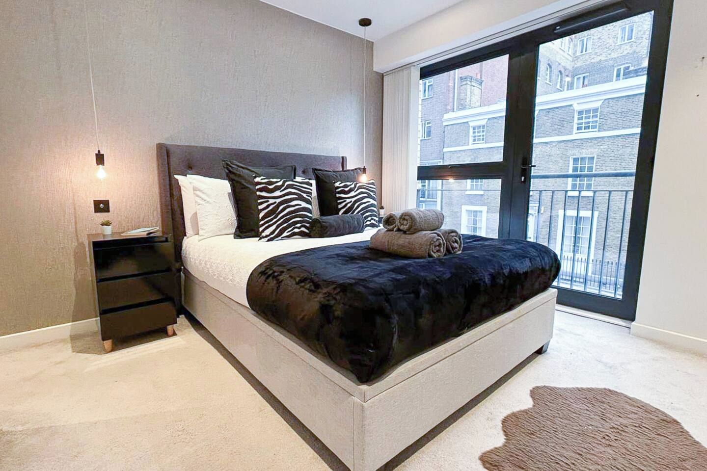 Ein modernes Schlafzimmer mit einem großen Bett, einem Nachttisch und Blick auf Gebäude durch ein großes Fenster.